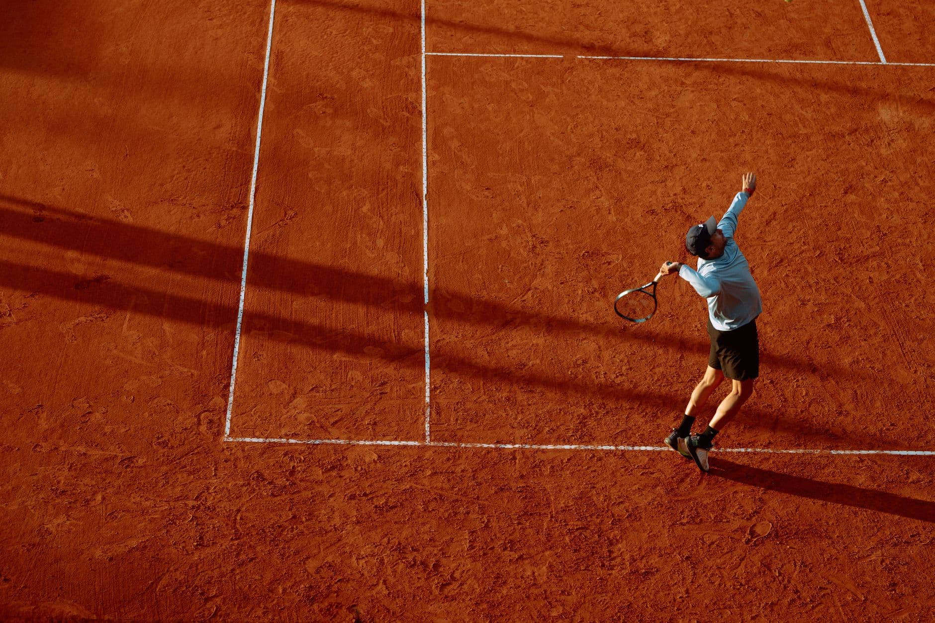 A tennis player serving on a clay court in low sun