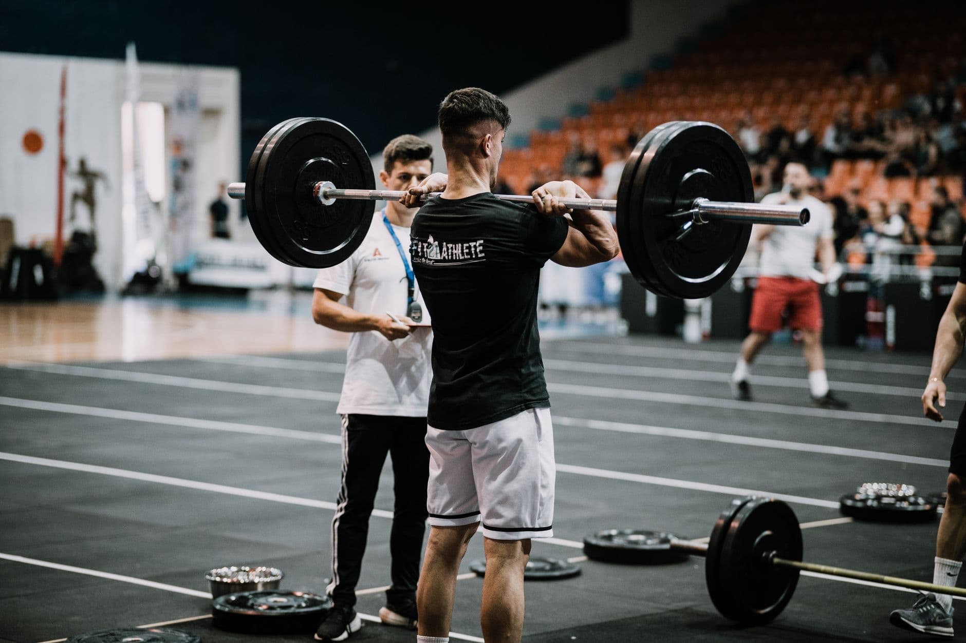 A lifter under the bar at a weightlifting competition, a judge watching